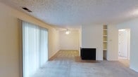 This single-floor space presents an open and functional layout centered around a combined living area. The room is finished with light-colored carpeting throughout, complemented by tile flooring at the fireplace hearth, which serves as a natural focal point. The fireplace features a dark surround, adding contrast to the otherwise neutral color palette of cream and beige tones that define the interior.

A ceiling fan is centrally positioned, providing both circulation and ambient light to the space. The textured ceiling adds subtle visual interest and is consistent throughout the room. To one side, built-in shelving units offer practical storage and display space, enhancing the room's functionality without requiring additional furniture.

Large sliding glass doors fitted with vertical blinds line one wall, allowing generous natural light to filter into the space and offering potential access to an outdoor area. The open layout between the living zone and the adjoining area creates a sense of spaciousness and easy flow. Overall, the space presents a clean, neutral canvas with practical built-in features well-suited for comfortable everyday living.