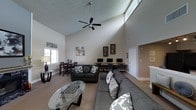 This single-floor space is defined by its impressive vaulted ceilings that rise dramatically overhead, creating an open and airy atmosphere throughout. A ceiling fan with a central light fixture hangs from the peak, providing both circulation and illumination to the expansive main area. Natural light filters in through strategically placed windows, including a high clerestory window near the roofline, brightening the neutral-toned walls and carpet.

The layout is thoughtfully organized into distinct functional zones. A generous lounge area anchors the space with two plush, dark gray sofas arranged around a glass-topped oval coffee table, offering a comfortable setting for relaxation or socializing. Adjacent to the lounge, a row of computer workstations lines a long desk, equipped with multiple monitors and task chairs — making it a practical resource for residents or visitors needing computer access.

A dining area with a dark wood table and matching chairs occupies a corner near the windows, providing an inviting spot for casual meals or informal meetings. Framed artwork decorates the walls, adding a touch of character to the space. A hallway visible in the background suggests additional rooms or amenities beyond the main area. Overall, the space balances comfort and functionality in a welcoming, communal environment.