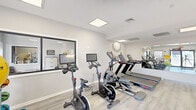 Located at 120 Estates Blvd in Trenton, NJ, this fitness center offers a well-appointed, single-floor exercise facility designed to support a comprehensive workout experience. The space features a clean, modern aesthetic with light gray walls, light wood-style flooring, and recessed panel lighting that creates a bright and energizing atmosphere throughout.

The cardio area is equipped with multiple treadmills, stationary bikes with integrated display screens, and an elliptical machine, all arranged to maximize usable floor space. Large wall-mounted mirrors extend the visual depth of the room while allowing users to monitor their form during exercise. A dedicated strength training area includes a tiered dumbbell rack with a full range of weights and an adjustable workout bench, ideal for resistance training routines.

Functional fitness accessories are also well-represented, with an assortment of medicine balls, stability balls, and resistance equipment neatly organized along the walls. A bulletin board and informational panels are mounted near the entrance, providing a practical touch for facility users.

Natural light filters in through windows flanking the main entrance, complementing the overhead lighting and contributing to an open, welcoming ambiance. The space benefits from a dedicated HVAC system and clearly marked exits, reflecting a thoughtful approach to both comfort and safety. Overall, this fitness center delivers a clean, fully equipped environment suited to a wide range of fitness goals.