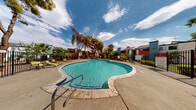 Located at 1900 East Tropicana Avenue in Las Vegas, Nevada, this outdoor amenity area centers around a generously sized, freeform swimming pool with a distinctive multi-lobed shape that adds a resort-style character to the space. The crystal-clear, turquoise water is complemented by a wide concrete pool deck that provides ample room for lounging and movement. Yellow lounge chairs are positioned along the deck, offering comfortable spots for sunbathing and relaxation.

The pool area is enclosed by a wrought iron fence, providing both security and an open, airy feel that preserves sightlines to the surrounding landscape. Mature trees — including tall palms and broad-canopy shade trees — frame the space, casting dappled shade and lending a lush, verdant atmosphere that softens the desert setting. Low-level plantings and shrubs add additional greenery throughout.

The surrounding residential buildings feature a Southwestern-influenced architectural style, with flat and sloped rooflines, stucco exteriors, and bold accent colors in red and blue. Pool entry is provided via built-in steps and handrails. The open sky above allows abundant natural light to flood the area throughout the day, creating a bright and inviting outdoor retreat well-suited to the warm Las Vegas climate.