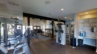 This fitness center is a spacious, single-floor facility designed to accommodate a comprehensive range of workout activities. The space is divided into distinct functional zones, each outfitted with dedicated equipment to support both cardio and strength training needs.

The main cardio area features a row of treadmills and elliptical machines positioned near a bank of windows that allow natural light to fill the room. Exposed dark structural beams run across the elevated ceiling in this section, adding an architectural contrast to the otherwise clean, light-toned walls. Durable, wood-look flooring underfoot provides a practical and visually appealing surface throughout this zone.

Adjacent to the cardio area, a dedicated strength training section houses a cable resistance machine, a multi-functional cable tower, and a rack system, offering versatile options for resistance-based workouts. An open floor space provides additional room for stretching, free-weight exercises, or functional training.

Recessed lighting supplements the natural light throughout, ensuring consistent illumination across all workout areas. The overall ambiance is clean and functional, with a neutral color palette of grey and white tones creating a focused, gym-appropriate environment.