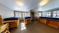 Located at 2315 College Ave in Berkeley, CA, this student residence room is situated on a single floor and designed to accommodate multiple occupants. The main living area is thoughtfully arranged with three lofted wooden bed frames, each paired with a matching dresser and built-in shelving unit, maximizing the use of available floor space. Multiple windows on two walls allow generous natural light to fill the carpeted room, creating a bright and functional environment throughout the day. A sliding-door closet with a hanging rod provides additional shared storage.

Each resident is equipped with a dedicated wooden desk and chair, complete with overhead shelving and individual task lighting — ideal for academic work. A compact refrigerator and microwave are conveniently integrated into the living space, offering basic in-room convenience. The neutral-toned walls and carpet contribute to a clean, practical atmosphere suited for study and rest.

The private bathroom features a wall-mounted sink with a mirrored medicine cabinet above, and a full bathtub and shower combination equipped with accessibility grab bars, a handheld showerhead, and tile flooring — reflecting a thoughtful, inclusive design. Overall, the space is efficiently organized to support the daily needs of student residents in a structured, communal living setting.