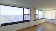 This single-floor space is defined by its expansive window wall, which stretches across the main living area and frames a broad, elevated panoramic view of the surrounding cityscape and tree-lined neighborhoods below. The generous glazing floods the interior with abundant natural light throughout the day, creating an airy, open atmosphere. Clean white walls and warm-toned hardwood flooring complement each other, contributing to the space's modern and minimalist character. Vertical blinds installed along the windows offer flexible light control while maintaining the visual connection to the outdoors. A built-in baseboard heating unit runs along the base of the window wall, ensuring comfort during cooler months without interrupting the room's clean lines. The open floor plan allows for versatile furniture arrangement, making the main living area adaptable to various uses. The elevated positioning of the space relative to its surroundings is a standout feature, offering unobstructed sightlines across rooftops and into the broader urban landscape. Overall, the space presents a bright, well-appointed interior with a strong emphasis on natural light and panoramic city views.