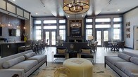 This single-floor space exudes upscale, contemporary design with a carefully curated interior that balances comfort and elegance. The room is anchored by a striking circular chandelier with candle-style lighting, suspended from a recessed ceiling that adds architectural depth. A dark-framed fireplace serves as the focal point of the far wall, flanked by tall, floor-to-ceiling windows and glass-paneled doors that flood the interior with natural light and provide views of the surrounding landscape. The dark wood trim framing the windows and doors lends a refined contrast to the lighter walls and ceiling. Seating is arranged thoughtfully throughout, including plush gray sofas, sleek accent chairs, and bar-height seating along the perimeter, catering to both relaxed lounging and more structured gatherings. A textured area rug anchors the central seating arrangement, which includes a distinctive gold ottoman as a stylish centerpiece. Decorative table lamps, glass accent tables, and carefully placed accessories contribute to the polished, layered aesthetic. The overall ambiance is warm yet sophisticated, making the space feel both inviting and visually impressive.