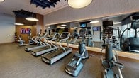 Located at 4850 Rugby Avenue in Bethesda, Maryland, this contemporary fitness center offers a well-appointed, single-floor layout designed to support a full range of workout routines. The space is characterized by an open, airy feel, enhanced by large street-facing windows that flood the cardio area with natural light and provide views of the surrounding neighborhood.

The cardio zone is generously outfitted with a row of Star Trac treadmills, elliptical trainers, and additional aerobic equipment, all positioned to take advantage of the natural light. Overhead, distinctive geometric acoustic panels in a dark finish add a modern architectural element to the ceiling, complemented by large circular pendant lights that provide warm, even illumination throughout.

The free weights area features a full rack of dumbbells alongside adjustable benches, all set against a floor-to-ceiling mirror that visually expands the space. A striking vertical wood slat partition serves as a refined architectural divider, adding warmth and texture to the otherwise sleek, neutral interior.

A dedicated functional training zone includes a wall-mounted display screen and a medicine ball rack, catering to varied fitness needs. Warm wood accents, textured wall finishes, and neutral-toned carpet throughout unify the space with a sophisticated, hotel-quality aesthetic.