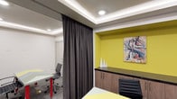 This single-floor space offers a thoughtfully designed interior that balances functionality with contemporary aesthetics. The layout incorporates flexible spatial division through a floor-to-ceiling charcoal curtain partition, allowing areas to be separated or opened as needed — an ideal feature for collaborative or private work settings.

A standout design element is the bold chartreuse yellow recessed niche, which serves as both a display and storage feature. Set against the otherwise neutral palette of white and gray tones, this vibrant accent wall adds a dynamic focal point to the space. Built-in cabinetry with a dark countertop runs beneath the niche, providing practical storage with a polished finish. Decorative white sculptural pieces and an abstract painting complete the vignette, lending an artistic sensibility to the environment.

Recessed LED lighting, including cove lighting integrated into a coffered ceiling detail, ensures even, ambient illumination throughout. The workstations are equipped with ergonomic task chairs in black and chrome, paired with light-toned work surfaces. The overall ambiance is professional yet creative, making the space well-suited for office or studio use.