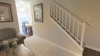 This two-story home is designed with a functional and open layout that connects its living spaces seamlessly. The entryway serves as the central hub of the main level, featuring plush wall-to-wall carpeting that transitions into warm hardwood flooring in the adjacent dining area. A white-railed staircase with clean balusters rises prominently from the entry, providing access to the upper level while contributing an airy, classic aesthetic to the space.

The neutral, warm-toned walls throughout create a cohesive and inviting atmosphere, complemented by white trim and baseboards that add a crisp, polished finish. A framed decorative mirror on the entryway wall adds depth and character to the space.

The dining area, visible from the entry, benefits from natural light filtering through a window with blinds, illuminating the hardwood floors and wooden dining set. An overhead light fixture provides additional ambiance. The overall design balances practicality with a warm, lived-in charm, making the space feel both comfortable and well-organized across both floors.
