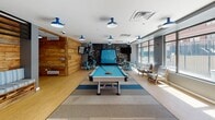This single-floor amenity space combines an urban-industrial aesthetic with playful, social design elements throughout its interconnected zones. Light hardwood flooring and a consistent palette of cool grays and blues unify the layout, while bold blue industrial-style pendant lights provide warm, focused illumination across all areas.

The main recreation area serves as the centerpiece, anchored by a Playcraft billiard table set atop a striped area rug. A dramatic chalkboard-painted accent wall featuring hand-lettered artwork and a painted van mural adds a creative, youthful energy to this zone, complemented by metal café-style chairs and a wall-mounted flat-screen television.

Adjacent to the recreation area, the entry and lounge corridor features a long built-in banquette upholstered in a striped fabric, set against a striking reclaimed wood accent wall. A service counter with blue metal bar stools serves as a central hub, framed by steel-and-glass partition windows that allow sightlines throughout.

A dedicated lounge and business center area showcases a bold geometric mural on a gray accent wall, alongside a comfortable sectional sofa, leather armchairs, and computer workstations. Large windows throughout the space flood the interiors with natural light and offer outdoor views, while a sunlit seating nook with paired lounge chairs and yellow ottomans provides a relaxed, informal retreat.
