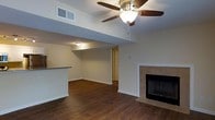 This single-story space showcases an open-concept layout that seamlessly connects the living area to the kitchen. The living room is anchored by rich, dark hardwood-style flooring that extends throughout, adding warmth and visual continuity to the space. A ceiling fan with a built-in light fixture is centrally mounted, providing both air circulation and ambient lighting. A tiled fireplace with a sleek black surround serves as a focal point along one wall, offering a cozy architectural detail.

A structural soffit defines the boundary between the living and dining or kitchen areas, where a partial countertop is visible, suggesting a breakfast bar or pass-through design. Neutral-toned walls throughout the space contribute to a clean, versatile aesthetic. Recessed HVAC vents are integrated into both the ceiling and walls, ensuring efficient climate control. A flush-mount ceiling light in the adjacent area provides additional illumination. White baseboards provide crisp contrast against the flooring, while a hallway visible in the background hints at further rooms beyond the main living space.