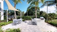 This outdoor patio space offers a well-appointed al fresco environment designed for relaxation and entertaining. The expansive paver-tiled terrace provides generous room for multiple seating arrangements, including sleek white dining tables and chairs that complement the space's clean, modern aesthetic.

A standout feature is the built-in outdoor kitchen, complete with a stainless steel grill and a white countertop island, making it ideal for outdoor cooking and casual gatherings. A shade sail stretched overhead offers a sheltered area for dining, providing relief from direct sunlight while maintaining an open-air feel.

The space is framed by tall, mature palm trees and lush tropical greenery, creating a resort-like ambiance. Neatly maintained shrubs and plantings line the perimeter, which is enclosed by a white metal fence, ensuring both privacy and a tidy boundary. The combination of durable paver flooring, tropical landscaping, and modern outdoor furnishings gives this patio a polished yet relaxed character, well-suited to a warm, sunny climate.