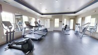 This fitness center offers a clean, well-organized environment designed to support a full range of workout routines. The space is finished with durable rubber flooring suited for both cardio and strength training activities. A row of treadmills lines one side of the room, accompanied by an elliptical machine, providing ample options for cardiovascular exercise. On the opposite side, a dumbbell rack and weight bench offer dedicated space for strength training. Large windows are positioned throughout the room, filling the space with natural light and contributing to an open, airy atmosphere. A full-length mirror mounted along one wall enhances the sense of depth while allowing users to monitor their form. The ceiling features a tray design with a contrasting navy border and recessed lighting, adding a polished architectural detail to the room. Neutral-toned walls complement the overall clean and functional aesthetic. The layout is thoughtfully arranged to allow comfortable movement between equipment, making the space accessible and practical for everyday use.