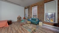 This single-floor loft showcases a compelling blend of industrial character and contemporary functionality. The living area is defined by striking exposed brick walls that span the length of the space, framing tall windows that allow generous natural light to fill the room. Hardwood-style flooring runs throughout, unifying the open-plan layout and lending warmth to the industrial aesthetic.

The living space comfortably accommodates a seating arrangement alongside a dedicated workspace, making it well-suited for both relaxation and productivity. A structural column adds to the loft's original architectural character. The open layout flows into a compact, efficiently designed kitchen finished in light tones. Pale blue walls contrast with warm wood flooring, while pendant cage lights hang from a tall ceiling above a continuous run of cabinetry and open shelving. The kitchen is equipped with a full-size refrigerator, dishwasher, electric range, and a single-basin sink.

The bathroom is straightforward and functional, featuring a wall-mounted sink, toilet, a shower with a curtain enclosure, and an above-mirror light fixture. The neutral gray walls create a clean, understated finish.