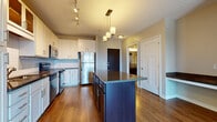 This single-floor residence showcases a thoughtfully designed open-concept layout centered around a well-equipped kitchen. The kitchen is a standout feature, offering white shaker-style cabinetry, a light blue subway tile backsplash, and a full suite of stainless steel appliances, including a refrigerator, electric range, built-in microwave, and dishwasher. A substantial dark-cabineted island with a granite countertop serves as a functional centerpiece, providing additional prep space and storage. Pendant lighting hangs above the island, complemented by track lighting overhead, creating a layered and well-lit environment. Warm hardwood flooring extends throughout the kitchen and main living areas, adding visual continuity and warmth. A built-in counter nook near the entry offers a convenient workspace or drop zone. The bedroom features neutral carpet, a ceiling fan, and a window that invites natural light and exterior views. The bathroom is finished with a granite-topped dark vanity, tile flooring, and a full tub-shower combination with chrome fixtures. The overall aesthetic is clean, modern, and functional, with consistent finishes throughout.