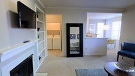 This single-floor residence is thoughtfully designed to maximize functionality while maintaining a clean, cohesive aesthetic throughout. The main living area is carpeted and features soft, neutral-toned walls that create a bright and airy atmosphere. A standout built-in shelving unit with white-painted woodwork lines one wall, offering generous display and storage space alongside a mounted flat-screen television. Crown molding adds a refined architectural detail throughout the space.

A full-length floor mirror with a bold black frame serves as both a practical and decorative focal point in the living area. Adjacent to the main living space, a dedicated laundry room houses a full-size washer and dryer, providing convenient in-unit laundry access. On the opposite side, an open pass-through connects to the kitchen, which benefits from natural light streaming through a window above the sink, creating a bright and welcoming cooking environment. The overall layout promotes an easy flow between living, utility, and kitchen areas, making the space both practical and comfortable for everyday living.