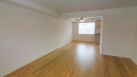 This two-floor space offers a clean and functional layout with notable architectural details. The main living area is generously proportioned, showcasing warm-toned hardwood floors that run throughout, lending a sense of continuity and natural warmth. Crisp white walls reflect light effectively, contributing to an open and airy feel. A structural beam accent is visible along the ceiling, adding a subtle architectural character to the space.

A ceiling fan with an integrated light fixture is centrally positioned, providing both ventilation and ambient lighting. The living area flows naturally toward a windowed section at the far end, where natural light filters in, brightening the interior. Adjacent to this area, a partially visible kitchen features cabinetry and counter space, connected to the main living zone in an open-concept arrangement.

The overall ambiance is understated and versatile, with neutral tones and hardwood flooring providing a solid foundation for a variety of interior styles. The two-floor configuration suggests additional living space beyond what is visible, offering ample room for diverse functional uses throughout the property.