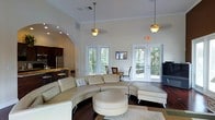 This single-floor space showcases a thoughtfully designed open-concept layout that seamlessly blends comfort and style. The main living area is a standout, featuring soaring ceilings adorned with crown molding and elegant pendant lights that cast a warm glow throughout. A sweeping curved sectional sofa anchors the room, complemented by a matching ottoman and a neutral color palette that enhances the sense of openness and light.

Large windows and multiple sets of French doors line the walls, flooding the interior with natural light and offering views of lush, tree-lined surroundings. The doors provide direct access to an outdoor area, further extending the living space and reinforcing the connection to nature.

A graceful arched passageway leads to a well-appointed kitchen, which features dark cabinetry, stainless steel appliances, and a raised bar counter with seating — ideal for casual dining or entertaining. The rich hardwood flooring flows consistently throughout, adding warmth and continuity to the overall design.

The space strikes a balance between airy, high-ceiling grandeur and intimate, functional living areas, making it both visually impressive and practically versatile. The overall ambiance is bright, welcoming, and refined, with architectural details that elevate every corner of the interior.