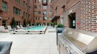 Located at 595 King Street in the heart of Charleston, South Carolina, this outdoor amenity space offers a well-appointed courtyard-style retreat set against the backdrop of a classic multi-story red brick building. The centerpiece of the space is a generously sized, rectangular swimming pool with clear turquoise water, shallow entry points, and a surrounding deck finished in large-format light stone tiles. Rows of cushioned lounge chairs are arranged along the pool's edge, providing ample space for sunbathing and relaxation.

Adjacent to the pool, a designated lounge area features comfortable all-weather wicker sofas and chairs arranged around a central focal point, ideal for outdoor socializing. A stainless steel outdoor grill station is conveniently positioned nearby, making the space well-suited for outdoor entertaining. Lush potted evergreen trees and tropical palms are strategically placed throughout the deck, adding greenery and a sense of privacy within the urban setting.

Warm pendant lighting and wall-mounted fixtures ensure the space is welcoming into the evening hours. The surrounding brick architecture frames the courtyard with a timeless, historic Charleston aesthetic, while the open-sky setting provides abundant natural light throughout the day. The overall ambiance is polished, functional, and inviting.