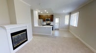 This single-floor residence offers a thoughtfully designed open-concept layout that seamlessly connects the living and kitchen areas. The living room is anchored by a classic white-framed fireplace with a sleek glass front, adding warmth and character to the space. Soft, neutral-toned carpeting flows throughout the living area, complementing the light gray walls and white crown molding that frame the room with a clean, polished finish.

The kitchen is a standout feature, showcasing rich dark wood cabinetry that contrasts beautifully with light countertops. Stainless steel appliances — including a refrigerator, microwave, and range — provide a modern, functional aesthetic. A peninsula with a built-in sink offers additional counter space and creates a natural divide between the kitchen and living areas, ideal for both everyday use and entertaining.

Recessed lighting throughout the ceiling ensures consistent, even illumination, while multiple large windows dressed with white blinds invite generous natural light into the space. A glass-panel door provides access to the exterior, further enhancing the bright, airy ambiance. The overall design strikes a balance between contemporary style and comfortable living, making this a welcoming and highly functional space.