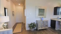 This single-floor space opens with a well-appointed entryway that sets a refined tone throughout. The entry corridor features rich, dark hardwood-style flooring that provides a striking contrast against the warm neutral walls painted in soft beige tones. Recessed lighting overhead illuminates the space efficiently, creating a bright and inviting atmosphere.

A decorative console cabinet with intricate scroll detailing serves as a focal point along the entryway wall, styled with a blue-and-white ceramic vase and complemented by a framed nature-inspired artwork above. A potted leafy plant adds a natural, organic element to the composition, enhancing the overall warmth of the space.

The entryway leads down a clean, well-proportioned hallway toward a paneled front door, which features a classic six-panel design. A woven area rug with a subtle border grounds the space near the entry, adding texture and visual interest underfoot. The clean lines, neutral color palette, and thoughtful decorative touches combine to create an entryway that feels both functional and aesthetically polished, offering an elegant first impression of the overall space.