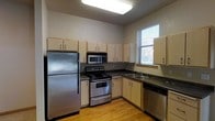 This single-floor space offers a practical and well-organized layout, highlighted by an open-concept design that connects the kitchen directly to the living area. The kitchen is fully equipped with stainless steel appliances, including a full-size refrigerator, an electric range with oven, a built-in microwave, and a dishwasher. Light wood-toned cabinetry lines the walls, providing generous storage space, while dark countertops offer a subtle contrast. A large flush-mounted ceiling light brightens the kitchen, supplemented by a window that introduces natural light and outdoor views. The flooring in the kitchen transitions from hardwood to carpet as it flows into the open living area, creating a clear visual distinction between the two spaces. The living area is finished with neutral-toned carpet and smooth walls painted in a warm beige, contributing to a clean and inviting atmosphere. Recessed lighting is present in the living space, adding to the overall brightness of the unit. A hallway extends toward additional rooms, suggesting further functional spaces beyond what is immediately visible. The overall design is straightforward and move-in ready.