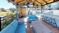 The Grilling Pavilion is a thoughtfully designed open-air structure that serves as a central hub for outdoor socializing and leisure. Anchored by a striking timber-framed roof with warm wood ceiling panels, the pavilion provides comfortable shade while maintaining an airy, open feel. A mounted flat-screen television is positioned beneath the peaked roofline, making it easy to enjoy entertainment from multiple seating areas.

The main covered deck features plush wicker lounge seating with vibrant blue cushions arranged around a central coffee table, creating a welcoming gathering space. A built-in bar counter with barstool seating lines one side of the pavilion, offering a convenient spot for casual dining and socializing. Stainless steel grills are stationed nearby, purpose-built for outdoor cooking.

The deck extends beyond the covered section, connecting seamlessly to a broader pool terrace furnished with additional dining sets shaded by green umbrellas. Natural wood-paneled accent walls add warmth and texture throughout the space. A wrought iron railing frames one edge of the pavilion, opening to serene views of a landscaped waterway and the surrounding community—enhancing the overall outdoor ambiance.