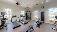 This fitness center is thoughtfully designed to support a comprehensive workout experience within a single, open-plan floor. The space is finished with durable speckled rubber flooring, neutral-toned walls, and recessed lighting complemented by ceiling fans, creating a comfortable and functional environment. Natural light pours in through multiple double-hung windows, offering views of the surrounding greenery and keeping the atmosphere bright and airy.

A full-wall mirror lines one side of the room, visually expanding the space and allowing users to monitor their form during exercise. The cardio section is well-appointed with multiple Matrix treadmills and an elliptical trainer, while a Matrix upright bike rounds out the aerobic equipment offerings. A multi-station cable machine serves as the centerpiece for strength and functional training, accompanied by a two-tier dumbbell rack stocked with a wide range of free weights and a flat workout bench.

A wall-mounted flat-screen television provides entertainment during workouts, and a fitness center rules board and sanitation station are conveniently positioned near the entry door. The space is accessed through a glass-paneled door, adding an elegant touch to an otherwise utilitarian setting. Overall, the fitness center strikes a balance between practicality and comfort, making it a well-rounded amenity for daily use.