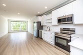 This single-floor unit presents a clean, contemporary design characterized by crisp white walls, light-toned hardwood flooring, and bold black-framed windows that appear throughout. The open layout offers a generous sense of space, with exposed metallic ductwork running along the ceilings in key areas, lending an industrial-modern aesthetic to the interior.

The kitchen is equipped with white shaker cabinetry, stainless steel appliances including a range and microwave, a gold-toned faucet at the sink, and open shelving, combining functionality with modern style. A long hallway connects the various rooms and features recessed lighting and a window at the far end that draws in natural light.

Living and sleeping areas benefit from large black-framed windows, including a striking floor-to-ceiling grid window that frames outdoor greenery views. Ceiling fans are present in select rooms. The bathrooms include a soaking tub with shower, white vanity cabinetry, and black hardware accents, with hardwood-style flooring extending throughout. A dedicated walk-in closet features multi-tiered wire shelving, providing practical storage. Overall, the space maintains a cohesive minimalist character with modern industrial touches.