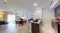 This single-floor residence showcases a thoughtfully designed open-concept layout that seamlessly connects the kitchen, dining, and living areas. Light-toned hardwood-style flooring extends throughout, creating a cohesive and airy aesthetic. The kitchen is equipped with stainless steel appliances, dark espresso cabinetry, granite countertops, and a peninsula island with bar seating — an ideal setup for both meal preparation and casual dining. Track lighting and recessed fixtures illuminate the space effectively.

The dining area accommodates a round table with seating for four, positioned centrally between the kitchen and living room. The living area is generously proportioned, featuring a ceiling fan with light fixture and sliding glass doors that open to an outdoor patio, allowing abundant natural light to fill the space.

The primary bedroom is well-sized, featuring a ceiling fan, large windows with patterned curtains, light hardwood-style flooring, and direct access to a private bathroom. A second bedroom offers comparable finishes, natural light, and ample space for furnishings. Both bedrooms maintain the clean, contemporary aesthetic found throughout the residence, contributing to an overall atmosphere that is bright, modern, and functional.
