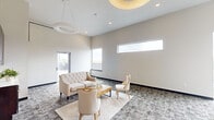 Located in Iowa City, Iowa, this thoughtfully designed hallway and lounge area offers a compelling blend of modern and warm design elements. The space is defined by its soaring ceilings, which lend an open, airy feel and amplify the sense of volume throughout. A striking floor-to-ceiling accent wall composed of mixed-tone wood planks in varying shades of brown, gray, and natural tones serves as the visual centerpiece of the space, adding texture and warmth against the otherwise neutral, light-colored walls.

Overhead, circular ring pendant lights provide stylish ambient illumination, complementing the recessed lighting integrated into the ceiling. Patterned carpet tiles in a muted gray tone cover the floors, adding a subtle visual interest underfoot. A built-in cabinetry unit with dark espresso-toned wood offers functional storage alongside a countertop, while a wall-mounted mini-split unit ensures climate comfort year-round.

The lounge area is furnished with cream-toned upholstered seating and a wooden coffee table, arranged to encourage relaxed gathering. Horizontal clerestory windows along one wall allow natural light to filter in without sacrificing wall space, while a glass door provides direct access to an outdoor terrace. A stainless steel elevator integrated into the accent wall adds a sleek, functional touch to this welcoming communal space.