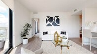 This contemporary single-floor residence is designed with a clean, modern aesthetic throughout, characterized by crisp white walls, light wood-tone flooring, and an overall bright and airy ambiance. The open-plan living area serves as the heart of the home, featuring a sleek seating arrangement anchored by a glass-top coffee table with gold accents and complemented by bold abstract wall art. The space flows seamlessly into a kitchen outfitted with a white breakfast bar, high-top seating, and stainless steel appliances. Adjacent to the living area, the dining space is distinguished by an elegant pendant light fixture with brass detailing, a glass-top dining table, and a gold-framed console, creating a refined yet approachable setting. Multiple bedrooms offer plush carpeting, generous walk-in closets, and access to private balconies with sweeping panoramic views through floor-to-ceiling sliding glass doors. Each bedroom maintains the home's signature neutral palette, with thoughtful built-in desk areas and ample dresser storage. The bathrooms are finished in light-toned tile with large vanity mirrors, undermount sinks, and full soaking tubs with shower enclosures, delivering a spa-like sense of calm. Throughout, the residence balances modern design with comfortable livability.