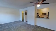 This single-floor residence offers a practical and comfortable layout designed for everyday living. The main living area is open and airy, finished with neutral-toned carpeting that adds warmth underfoot. A ceiling fan with an integrated light fixture is centrally positioned overhead, providing both ventilation and ambient lighting to the space. Clean white walls throughout contribute to a bright, welcoming atmosphere.

The kitchen is partially open to the living area, separated by a half-wall that doubles as a breakfast bar with a granite-style countertop. Upper cabinetry in a light finish provides ample storage, while a dark-toned backsplash adds a subtle contrast to the kitchen design.

From the living area, an open corridor leads to additional spaces, including a bedroom and a spacious walk-in closet visible through an open doorway. A second doorway with a dark-framed door is also accessible from this hallway, suggesting a bathroom or additional room beyond. The layout flows naturally, making efficient use of the available square footage while maintaining a sense of openness throughout. Overall, the space presents a clean, functional, and move-in-ready environment suited for comfortable residential living.