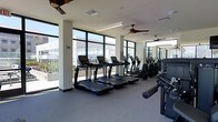 This single-floor fitness center offers a bright and spacious workout environment designed for an effective exercise experience. Floor-to-ceiling windows line the perimeter, flooding the space with abundant natural light while providing open views of the surrounding exterior landscape, creating an energizing atmosphere for workouts.

The cardio zone features a row of treadmills and elliptical machines equipped with individual digital screens, allowing users to track performance metrics during their sessions. Ceiling fans are mounted throughout, promoting comfortable air circulation across the workout floor. The neutral-toned walls and light gray flooring give the space a clean, contemporary aesthetic.

Strength training equipment is positioned along one side of the room, with a mirrored wall extending behind it to enhance the sense of depth and allow users to monitor their form. The layout is thoughtfully organized to ensure clear separation between cardio and strength areas, facilitating smooth traffic flow. The combination of modern equipment, ample natural light, and a well-planned open layout makes this fitness center a functional and inviting space for users of all fitness levels.