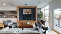 This thoughtfully designed single-floor space blends modern sophistication with warm, natural materials to create an inviting lounge and entertaining environment. The open layout flows seamlessly between a comfortable living area and a functional wet bar and dining zone, all grounded by wide-plank hardwood flooring that runs throughout.

The living area serves as the heart of the space, anchored by a striking feature wall that frames a large flat-screen television above a sleek built-in fireplace. The surround is finished in dark shiplap-style paneling set within a bold natural wood frame, creating a strong focal point. Generous windows flood the space with natural light and offer elevated outdoor views, extending to an accessible balcony or terrace beyond.

The wet bar area is equally well-appointed, featuring a grey subway tile backsplash, white cabinetry with brass hardware, open wood shelving for display and storage, a sink, and an under-counter beverage refrigerator. Pendant lights with brass and wood finishes hang above a rustic reclaimed-wood dining table surrounded by industrial-style swivel stools, completing the casual yet refined aesthetic.

Glass-paneled doors connect the main space to an adjacent room, maintaining visual openness while allowing for separation. Clean white walls, recessed lighting, and carefully curated decor contribute to an overall ambiance that is warm, modern, and well-suited for both relaxed gatherings and social entertaining.