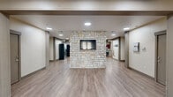 Located at 100 Hawk Ridge Dr in Iowa City, Iowa, this generously sized single-floor community amenity space offers a well-rounded mix of social, recreational, and functional areas. The open layout is anchored by a striking natural stone accent wall with a wall-mounted flat-screen TV, serving as a central visual focal point that connects the various zones of the space.

A standout feature is the dramatic game room, enclosed by floor-to-ceiling windows that flood the area with natural light and provide sweeping views of the surrounding landscape. This bright room accommodates a full-size pool table and ping-pong table, making it an ideal recreational hub. Stone wainscoting lines the lower walls, adding continuity to the overall design aesthetic.

The lounge area features a stone-clad fireplace with a traditional mantle, double-sided visibility, and soaring double-height ceilings with an upper-level balcony overlooking the space. A mounted flat-screen TV and oval dining table with industrial-style chairs complete this inviting gathering zone.

The kitchen is fully equipped with rich cherry wood cabinetry, granite countertops, a stainless steel refrigerator, double-basin sink, and a stone-accented island with bar seating — ideal for events and communal dining. A secondary prep kitchen provides additional counter space and storage. Throughout, light wood-tone flooring ties the entire space together with a clean, modern finish.