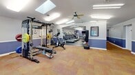 Located at 1712 Waverly Way in Baltimore, MD, this fitness center offers a generously sized, single-floor exercise space designed to support a comprehensive workout experience. The facility is divided into distinct functional zones, seamlessly connected through an open archway that creates a sense of expansive flow throughout the room.

The strength training area features a Major Fitness power rack loaded with weight plates, adjustable benches, dumbbells, and a Hoist cable machine, providing a full range of resistance training options. A heavy bag rounds out the strength zone for additional training variety.

The cardio section is well-appointed with multiple Life Fitness treadmills, elliptical cross-trainers, and upright stationary bikes, each equipped with individual digital displays. Durable rubber flooring in this area provides cushioning and stability during high-impact workouts.

Architectural highlights include a vaulted ceiling with multiple skylights that flood the space with natural light, complemented by ceiling fans and fluorescent panel lighting for year-round comfort. The walls feature a two-tone color scheme with a periwinkle blue accent band, adding a clean, motivating aesthetic. A wall-mounted television is centrally positioned for entertainment during workouts. Carpeted flooring in the free weights zone and direct exterior access via glass-panel doors complete this practical and inviting fitness facility.