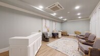 This thoughtfully designed beauty salon or spa suite offers a refined, single-floor layout that balances professional functionality with a calm, welcoming ambiance. The space is finished throughout in soft sage-gray tones, complemented by light hardwood-style flooring that adds warmth and continuity across every area.

At the entrance, a crisp white paneled reception desk serves as the focal point, accented by recessed lighting set within a subtly coffered ceiling. A decorative area rug with soft, muted tones anchors the central floor space, adding texture and visual interest. Abstract artwork in warm neutral hues adorns the walls, contributing to the space's polished yet approachable character.

The main salon area features multiple styling stations, including a dedicated shampoo bowl paired with a tan reclining chair, a professional styling chair beside a white dresser with a framed mirror, and a manicure table with client seating. Toward the window wall, sheer white curtains diffuse soft natural light, illuminating a seating area furnished with elegant French-style upholstered chairs arranged around the central rug.

Crown molding details and a ceiling-mounted HVAC unit reflect careful attention to both aesthetics and comfort, making this a highly functional, beautifully finished professional environment.