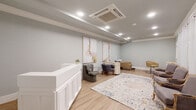This thoughtfully designed beauty salon or spa suite offers a refined, single-floor layout that balances professional functionality with a calm, welcoming ambiance. The space is finished throughout in soft sage-gray tones, complemented by light hardwood-style flooring that adds warmth and continuity across every area.

At the entrance, a crisp white paneled reception desk serves as the focal point, accented by recessed lighting set within a subtly coffered ceiling. A decorative area rug with soft, muted tones anchors the central floor space, adding texture and visual interest. Abstract artwork in warm neutral hues adorns the walls, contributing to the space's polished yet approachable character.

The main salon area features multiple styling stations, including a dedicated shampoo bowl paired with a tan reclining chair, a professional styling chair beside a white dresser with a framed mirror, and a manicure table with client seating. Toward the window wall, sheer white curtains diffuse soft natural light, illuminating a seating area furnished with elegant French-style upholstered chairs arranged around the central rug.

Crown molding details and a ceiling-mounted HVAC unit reflect careful attention to both aesthetics and comfort, making this a highly functional, beautifully finished professional environment.