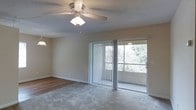 This single-floor space centers around a spacious living area that blends comfort and functionality. The room features neutral-toned walls and a combination of carpet and hard flooring, creating a warm and inviting atmosphere. A ceiling fan with an integrated light fixture is mounted on the textured ceiling, providing both air circulation and ambient lighting throughout the space.

A prominent sliding glass door, framed by vertical blinds, serves as the focal point of the main living area. It allows an abundance of natural light to fill the room while offering direct access to an outdoor area, where mature greenery is visible, adding a refreshing natural backdrop. The large adjacent window further enhances the brightness of the space, making it feel open and airy.

The transition between the carpeted living area and the hard surface flooring defines distinct functional zones within the room. Neutral wall tones throughout provide a versatile canvas suitable for a variety of interior styles. The overall ambiance is bright and livable, with practical features that enhance day-to-day comfort.