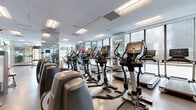 Located at 100 Park Ave in Fort Lee, New Jersey, this well-appointed fitness center offers a spacious, single-floor layout designed to accommodate a full range of workout activities. The facility features a clean, contemporary aesthetic characterized by a neutral color palette, smooth flooring, and a drop ceiling fitted with recessed panel lighting that ensures consistent, even illumination throughout.

Floor-to-ceiling glass windows line the perimeter, flooding the space with abundant natural light and offering open views of the surrounding Fort Lee skyline. Full-length mirrors are strategically positioned along the walls, enhancing the sense of openness and allowing users to monitor their form during exercise.

The cardio zone is well-equipped with multiple treadmills, elliptical trainers, and stationary bikes — all featuring modern digital consoles. Strength training is supported by a Synrgy360 functional training system, a selection of resistance machines including a leg press, and an array of stability balls for functional fitness. The layout is thoughtfully organized, providing clear circulation paths between equipment zones.

Glass partitions divide sections of the facility, adding a modern architectural touch while maintaining a sense of visual continuity. Overall, the fitness center presents a bright, professional, and fully equipped environment suited for a broad range of fitness goals.