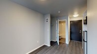 This single-floor unit offers a clean, contemporary design with a practical and efficient layout. The main living area features dark hardwood-style flooring that contrasts elegantly with light grey walls, creating a modern and neutral aesthetic throughout. Recessed and flush-mount ceiling lighting ensures consistent illumination across the space.

An open closet area with a built-in rod provides convenient storage directly off the main room, while a full private bathroom is accessible through a white-framed door. The bathroom is well-appointed with a bathtub and shower combination, toilet, and vanity sink, all finished in bright, clean tones that complement the overall style of the unit.

A dark-toned entry door provides a bold contrast to the lighter interior palette, contributing to the modern character of the space. A wall-mounted panel, likely for climate or intercom control, adds a functional touch near the closet area. White baseboards throughout give the unit a crisp, finished look.

The layout is straightforward and thoughtfully organized, making efficient use of space while maintaining an open, uncluttered feel. The consistent use of neutral tones and contemporary materials gives this unit a cohesive and move-in-ready appearance.