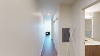 This single-floor unit offers a thoughtfully designed layout that maximizes functionality within a compact footprint. Wood-look flooring runs continuously throughout the space, lending warmth and visual cohesion to every room.

The entry corridor is clean and minimalist, lined with white walls and illuminated by recessed ceiling lighting. A wall-mounted electrical panel and a freestanding coat rack are positioned near the entrance, providing practical organization from the moment you step inside. A sliding door along the hallway leads to additional areas of the unit.

The living area is a standout feature, anchored by a large floor-to-ceiling window that floods the room with natural light and frames open views of the surrounding neighborhood and treetops. The space comfortably accommodates a seating area and a dedicated workspace, making it well-suited for both relaxation and productivity.

Adjacent to the living area, the kitchen is efficiently arranged in an L-shaped configuration. It features stainless steel appliances—including a refrigerator, electric range, and over-the-range microwave—paired with two-tone cabinetry combining white upper cabinets and warm wood-finish lower drawers, light countertops, and a single-basin sink.

The bedroom is a quiet, simply finished room with neutral walls. The full bathroom includes wood-finish cabinetry, a combined tub and shower, and wood-look flooring consistent with the rest of the unit.