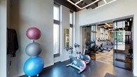 This contemporary fitness center is designed to provide an energizing and well-equipped workout environment. The space is immediately distinguished by its dramatic double-height ceilings, which create an open, airy atmosphere that enhances the sense of space throughout. Large floor-to-ceiling windows line the walls, flooding the interior with natural light and offering views of the surrounding exterior, contributing to a bright and motivating ambiance.

The flooring consists of dark, durable material well-suited for high-traffic fitness use. The cardio zone is prominently featured, with a row of stationary cycling bikes positioned near the windows, allowing users to exercise with natural light. A mirrored wall section visually expands the space while reflecting additional cardio equipment, including treadmills, visible in the background. A set of stability and exercise balls in varying sizes is neatly stacked along one wall, adding a functional and colorful element to the room.

Overhead, geometric linear lighting fixtures are integrated into the ceiling design, providing consistent, modern illumination throughout. The overall design balances functionality with a clean, upscale aesthetic.