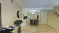 This single-floor residence offers a comfortable and cohesive living environment defined by an open, flowing layout that connects the main living and dining areas seamlessly. The dining space serves as a central focal point, featuring a dark wood dining table and chairs set beneath a stylish pendant light. A decorative circular wall accent adds a modern touch to the sage green accent wall, creating a warm and inviting atmosphere. Light-toned tile flooring runs throughout the main areas, contributing to a bright and airy feel. The entryway is accented with a round mirror and a lush indoor plant, lending a welcoming first impression. Adjacent to the dining area, the kitchen is partially visible and features white cabinetry, dark countertops, and standard appliances, offering a functional and tidy cooking space. The textured ceiling adds a classic residential character, while the neutral wall tones provide a versatile backdrop throughout. Natural light filters into the space, enhancing the overall warmth of the interior. The layout is practical and efficient, making thoughtful use of the available space to create distinct yet connected zones for dining, cooking, and everyday living.