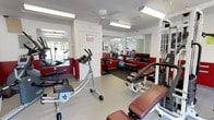 This fitness center is designed to accommodate a comprehensive workout experience within a single-floor layout. The space is immediately defined by its striking two-tone wall treatment, featuring bold red lower panels contrasted against white upper walls, creating an energetic and motivating atmosphere throughout.

The facility is well-lit by a combination of overhead fluorescent fixtures and abundant natural light streaming through multiple windows, including a large sliding glass door that frames views of the surrounding outdoor greenery. A ceiling fan provides additional air circulation in the main workout area.

Large wall-mounted mirrors are strategically placed throughout the space, enhancing the sense of openness and allowing users to monitor their form during exercise. The flooring transitions between smooth tile in some zones and rubber matting in designated workout areas, offering both durability and comfort.

The equipment selection is varied and well-suited for full-body training. Cardio options include a treadmill, elliptical trainer, and recumbent-style exercise bikes. Strength training is supported by a multi-station cable machine, adjustable benches, a dumbbell rack, and a stability ball. A water cooler and waste bin are conveniently positioned near the exit. Security is maintained via CCTV monitoring, as noted by posted signage.