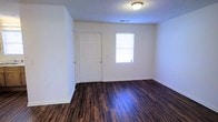 This single-floor space offers a straightforward and functional layout with a clean, neutral aesthetic throughout. The main living area features rich, dark hardwood-style flooring that contrasts sharply with the bright white walls and white baseboards, creating a crisp, well-defined interior. A paneled front entry door with a gold-tone handle serves as the main point of entry, positioned alongside a window that allows natural light to filter into the space, brightening the room and contributing to an open, airy feel. The ceiling features a textured finish and is equipped with a flush-mount light fixture, providing overhead illumination throughout the area. HVAC ventilation is visible in the ceiling, indicating climate control throughout the space. The walls are clean and unadorned, offering a blank canvas suitable for a variety of uses and décor styles. The overall ambiance is simple and understated, with practical finishes that are easy to maintain. The space presents a functional foundation ideal for those seeking a no-frills environment with durable flooring and a bright, open interior.