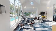 Located at 851 Church St in Mountain View, California, this two-story fitness facility combines thoughtful architecture with functional amenity spaces. The ground floor houses a generously sized fitness center defined by soaring vaulted ceilings, round clerestory windows, and an expansive wall of floor-to-ceiling windows that flood the room with natural light and offer views of an outdoor pool and lush greenery. Pendant globe lights complement the bright, airy atmosphere. The gym is fully equipped with cardio machines including ellipticals, stationary bikes, and treadmills, alongside a comprehensive selection of strength-training equipment and free weights, all arranged on a patterned blue modular carpet. A warm wood-paneled staircase with a light wood railing leads to the upper level. The second floor presents a spacious, open-plan studio with polished golden terrazzo floors that reflect the natural light streaming in through arched and vertical windows. The clean white walls, angled roofline, and sleek linear ceiling lighting give the upper level a modern, minimalist character, making it a flexible space ideal for yoga, group fitness classes, or meetings. A small open shelving unit near the staircase landing provides convenient storage. Together, the two floors offer a cohesive, light-filled environment designed to support a wide range of fitness and wellness activities.