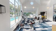 Located at 851 Church St in Mountain View, California, this two-story fitness facility combines thoughtful architecture with functional amenity spaces. The ground floor houses a generously sized fitness center defined by soaring vaulted ceilings, round clerestory windows, and an expansive wall of floor-to-ceiling windows that flood the room with natural light and offer views of an outdoor pool and lush greenery. Pendant globe lights complement the bright, airy atmosphere. The gym is fully equipped with cardio machines including ellipticals, stationary bikes, and treadmills, alongside a comprehensive selection of strength-training equipment and free weights, all arranged on a patterned blue modular carpet. A warm wood-paneled staircase with a light wood railing leads to the upper level. The second floor presents a spacious, open-plan studio with polished golden terrazzo floors that reflect the natural light streaming in through arched and vertical windows. The clean white walls, angled roofline, and sleek linear ceiling lighting give the upper level a modern, minimalist character, making it a flexible space ideal for yoga, group fitness classes, or meetings. A small open shelving unit near the staircase landing provides convenient storage. Together, the two floors offer a cohesive, light-filled environment designed to support a wide range of fitness and wellness activities.