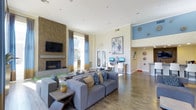 This single-floor space is a well-appointed communal lounge designed to blend comfort and functionality. The room is anchored by a striking floor-to-ceiling tiled fireplace wall, which serves as a focal point and adds texture and warmth to the overall aesthetic. A wall-mounted flat-screen television sits above the fireplace, enhancing the space's entertainment value. The seating area features generous gray sectional sofas adorned with patterned and neutral throw pillows, arranged to encourage social interaction.

Natural light flows in through large windows and French doors dressed with two-tone blue and brown curtains, creating a bright and welcoming atmosphere. The flooring throughout consists of warm wood-tone planks that complement the neutral cream and blue color palette of the walls. A built-in computer workstation with monitors and white chairs is conveniently positioned along one wall, offering a dedicated area for productivity. An adjacent alcove features blue accent walls, decorative wall plates, and a counter with bar-height seating, adding a casual dining or social nook to the space. The high ceilings contribute to an airy, open ambiance throughout.