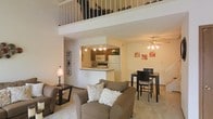 This two-story residence offers a well-designed open-concept layout that seamlessly connects the main living areas. The living room serves as the heart of the main floor, featuring a ceiling fan with light fixture, neutral carpeting, and ample space for comfortable seating arrangements. A loft overlooks the living area from above, framed by a white-painted balcony railing that adds architectural interest and a sense of vertical openness to the space.

The kitchen is positioned just off the living area and is accessible through a pass-through opening, creating a connected yet defined cooking space. It is equipped with standard appliances including a white refrigerator, microwave, and stovetop, along with dark wood cabinetry and track lighting overhead. A bar-height counter at the pass-through provides a convenient serving or casual dining surface.

Adjacent to the living room, the dining area accommodates a high-top table and chairs, making it ideal for everyday meals or entertaining. The main level flows naturally throughout, with consistent neutral tones in the walls and flooring that create a warm, cohesive ambiance. The interior staircase leads to the upper loft level, offering additional flexible living space that overlooks the main floor below.