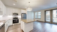 Located in Austin, TX 78757, this well-appointed one-bedroom, one-bathroom unit offers a thoughtfully designed single-floor layout with an efficient use of space. The open-concept kitchen and living area serves as the heart of the home, featuring crisp white cabinetry with brushed nickel hardware, a subway tile backsplash, quartz countertops, and a central island with pendant lighting overhead. A full suite of stainless steel appliances — including a refrigerator, range, microwave, and dishwasher — rounds out the kitchen's functional appeal. Wood-look plank flooring flows seamlessly throughout the living areas, lending warmth and continuity to the space. The living room is bathed in natural light through large windows and a set of glass doors that open to an outdoor view, creating an airy, open atmosphere. The spacious primary bedroom features a ceiling fan, generous natural light from wide windows, and the same hardwood-style flooring. The bathroom is finished in a clean, all-white palette with large-format tile floors, a soaking tub with a tiled surround, a single-sink vanity with quartz countertop, and open shelving for added storage. The unit's modern, neutral aesthetic makes it a versatile and inviting living space.